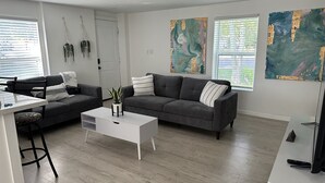 Een moderne woonkamer met een grijze bank, een witte salontafel en een kleine plant. Op de muren hangen abstracte schilderijen en er is een raam met zonwering.