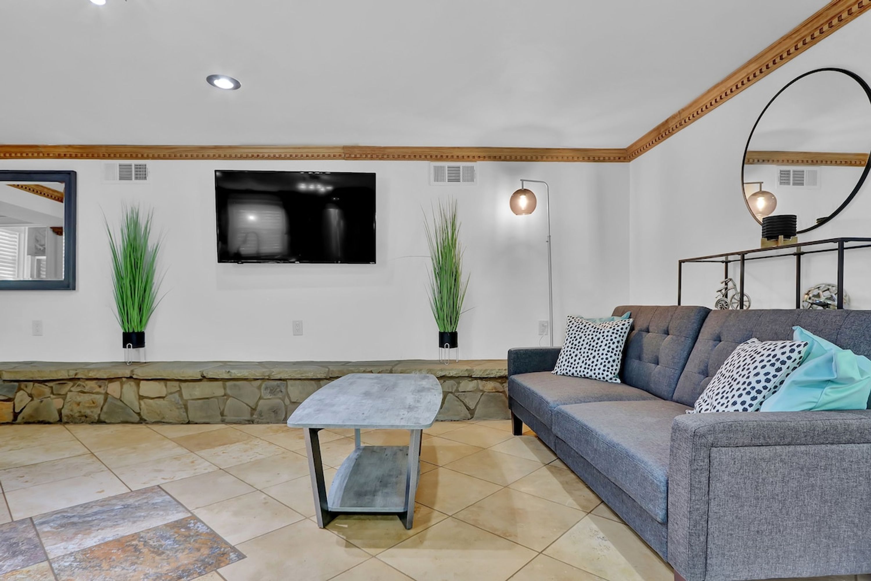 A living room with a grey sofa, a small table, a TV mounted on the wall, and two potted plants.