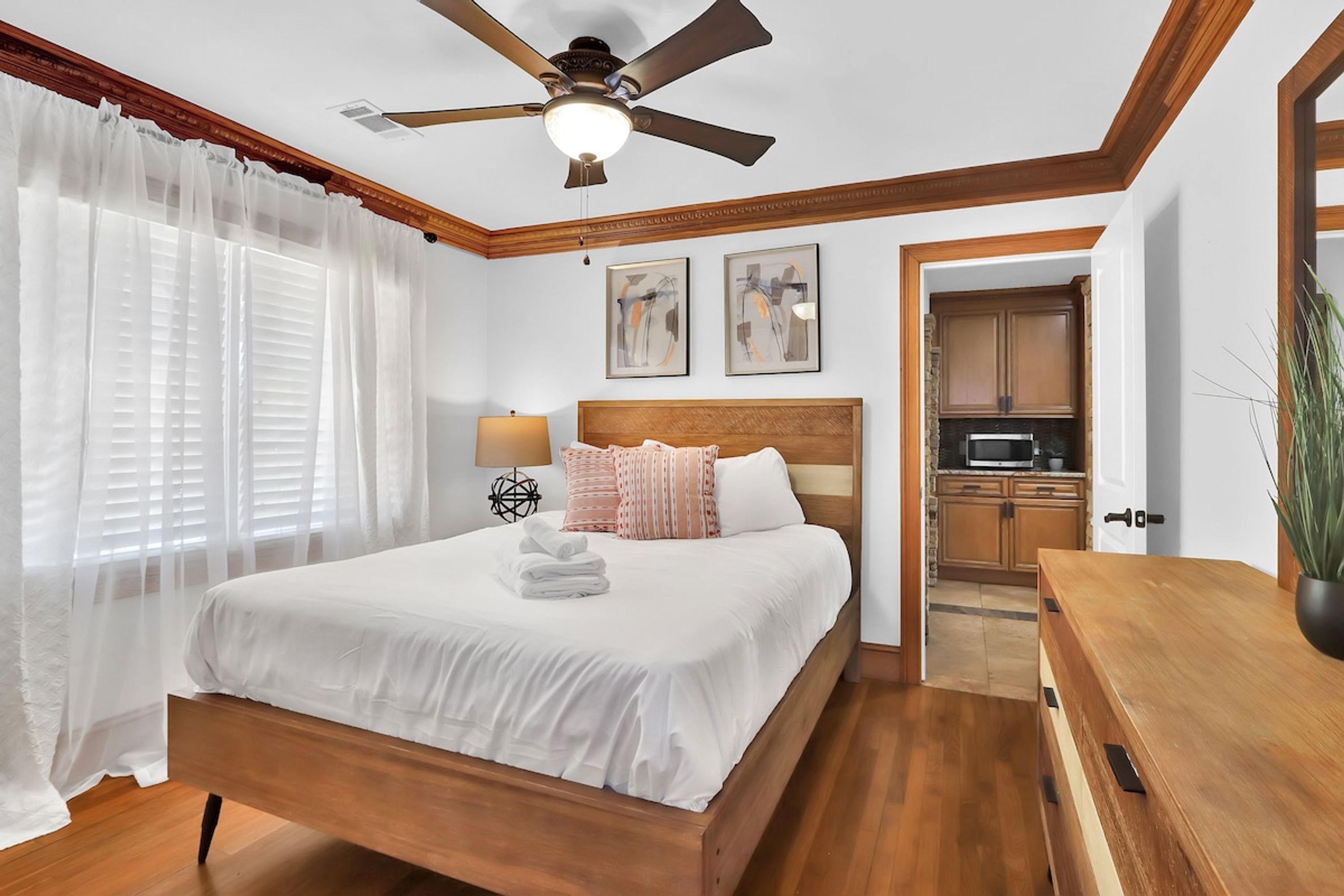 A bedroom with a wooden bed, white bedding, a ceiling fan, and a view into the kitchen.