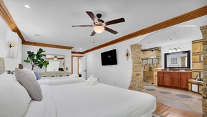 Camera d'albergo con due letti, una ventola a soffitto, una TV, un muro decorativo in pietra e un angolo cottura con mobili in legno.