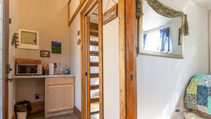 A cozy room with a wooden door, a welcome sign, a bed with a patterned quilt, a small kitchenette with a microwave, and a window with curtains.