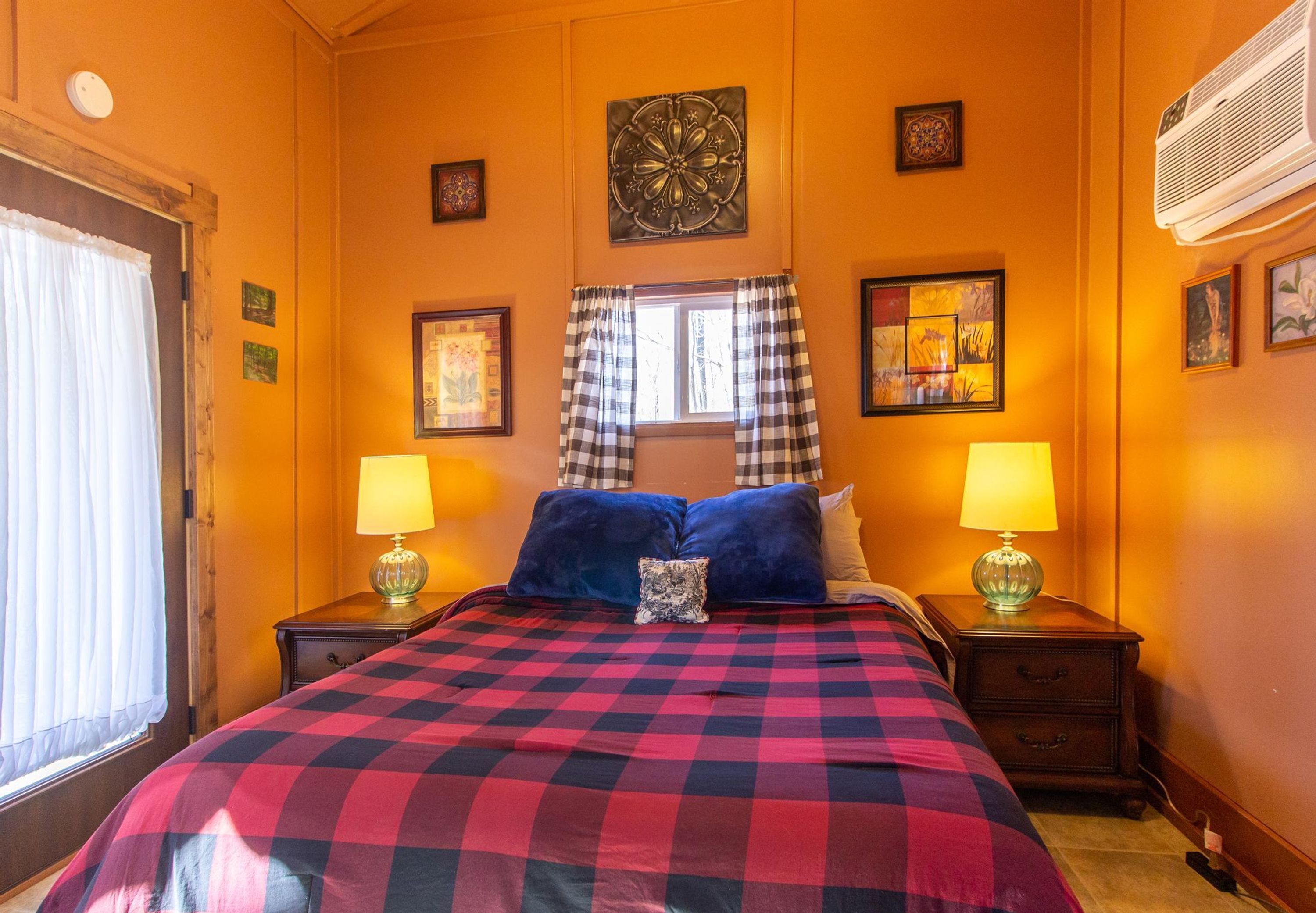 Een slaapkamer met een geruit beddegoed, twee nachtkastjes met lampen, een raam met geruiten gordijnen en wanddecoratie.