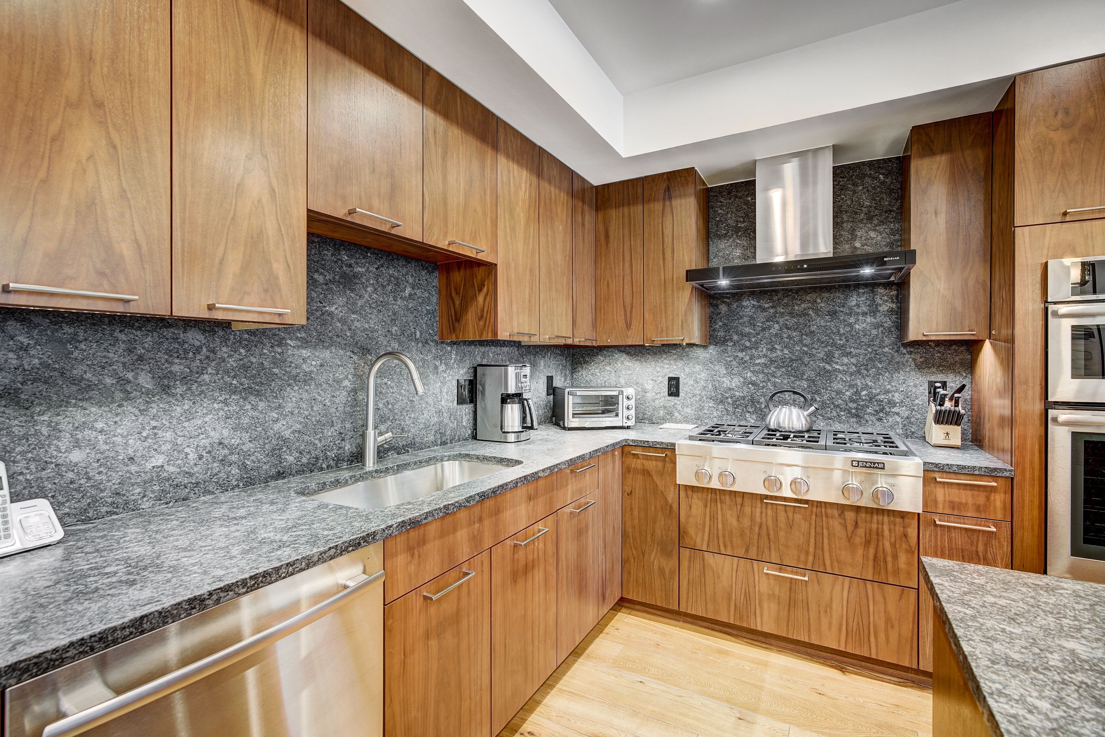 A modern kitchen with wooden cabinets, granite countertops, a stainless steel range hood, and a built-in oven.