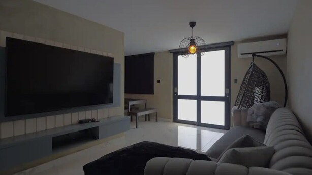 Een moderne woonkamer met een grote flatscreen televisie, een bank, een hanglamp en een schuifdeur naar een balkon.