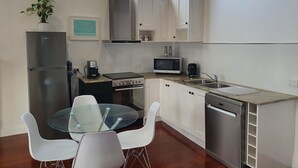 Une cuisine moderne avec des meubles blancs, des appareils en acier inoxydable, une table à manger en verre et une vue sur les arbres par la fenêtre.