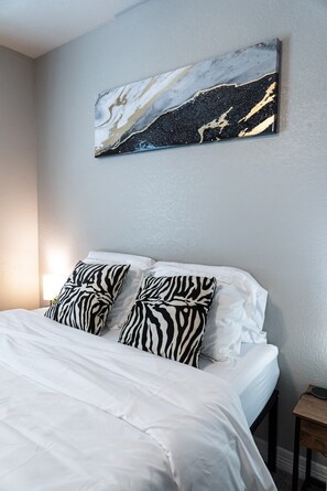 Un lit bien fait, avec une literie blanche et des coussins à motifs zébrés, une œuvre d’art abstraite accrochée au mur, et une petite table de chevet en bois avec une lampe.