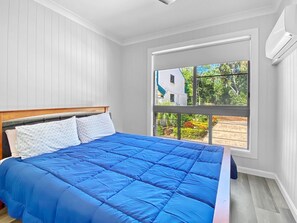 A bedroom with a large bed, a wooden headboard, a window with a view of greenery and a building, and a white wall. - Magnetic Reef Unit 2 (Arcadia)