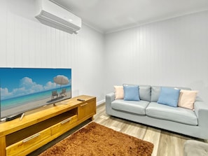 A modern living room with a flat-screen TV, a grey sofa with blue and beige cushions, a wooden TV stand, and a brown rug. - Magnetic Reef Unit 2 (Arcadia)