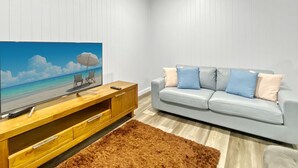 Een moderne woonkamer met een flatscreen televisie, een grijze bank met blauwe en beige kussens, een houten tv-meubel en een bruin vloerkleed.