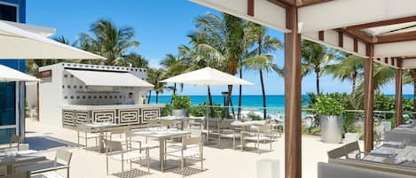 An outdoor dining area with tables and chairs, a bar, and a view of the beach and ocean.