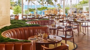 Un ristorante con tavoli rotondi e panche in pelle, che offre vista su palme e oceano.