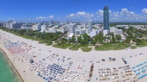Paesaggio urbano sul lungomare con un grattacielo in evidenza, una spiaggia di sabbia ampia con numerosi ombrelloni e un cielo azzurro terso.