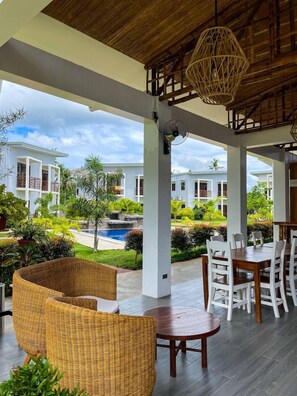 A spacious outdoor area with wicker furniture, a dining table, and a view of a swimming pool and buildings in the background. - Red Picnic Hostel (Panglao)