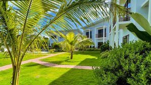 Un jardin bien entretenu, agrémenté de palmiers et d’un sentier menant à un bâtiment moderne doté de balcons.