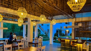 Un restaurant avec des tables et des chaises en bois, des luminaires suspendus et une vue sur l’extérieur.