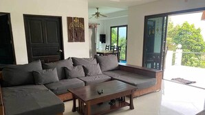 A modern living room with a sectional sofa, a wooden coffee table, and a glass door leading to a balcony. - Penthouse Seaview Apartment - 3 Bedroom, Sleeps up to 8 (Tambon Ko Tao)
