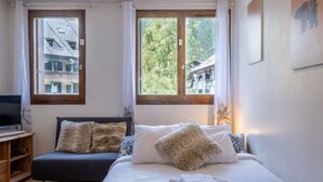 Une chambre à coucher avec un lit, un canapé, une télévision et des fenêtres donnant sur des immeubles et des arbres.