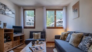 Un salon confortable avec un canapé, une table basse et un téléviseur. Il y a des fenêtres avec des rideaux, un meuble en bois et un tableau accroché au mur.