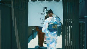 Une personne vêtue d’un costume traditionnel traverse un seuil, et une enseigne au-dessus indique « NIPPON ».