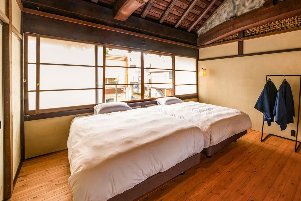A traditional Japanese-style bedroom with a wooden floor, a large window, a bed with white bedding, and a wall-mounted air conditioner.