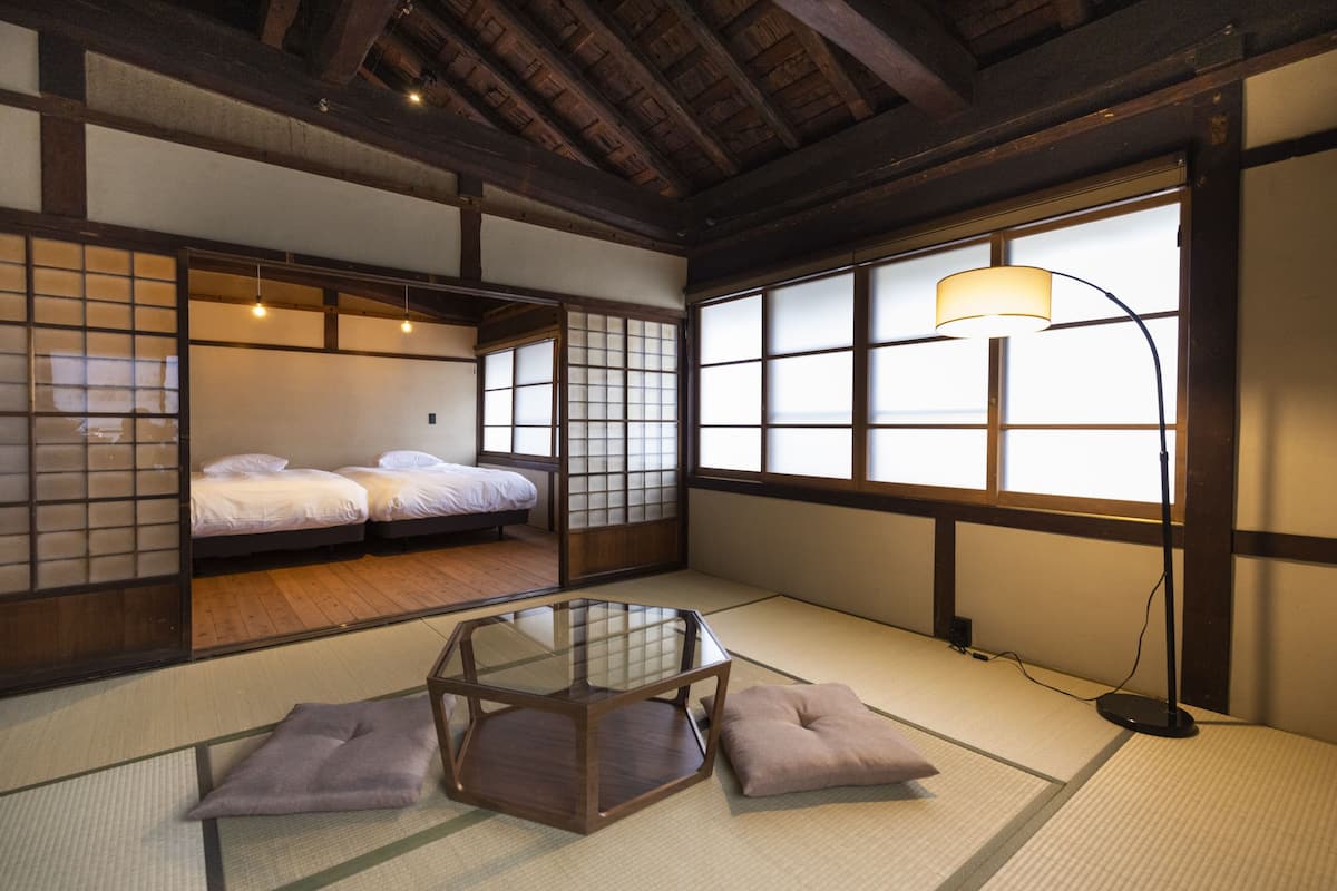 A traditional Japanese room with tatami flooring, a low table, a bed with futon beds, and a large window with a lamp.