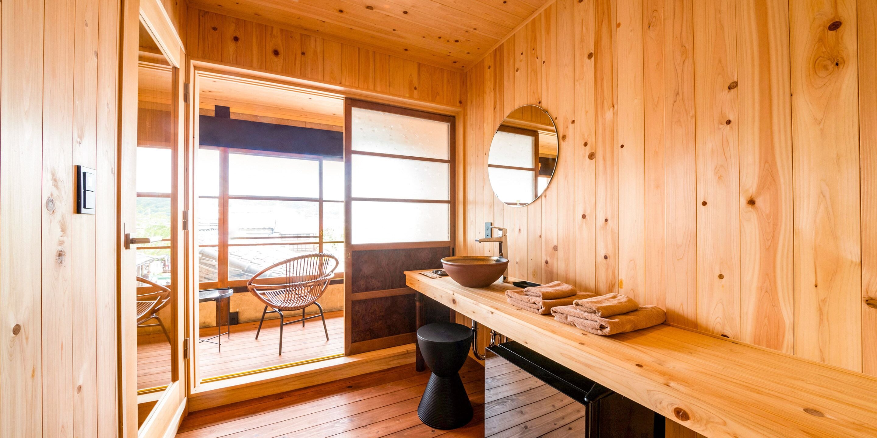 A wooden interior with a large sliding door leading to an outdoor area, a round mirror on the wall, and a wooden bench with towels.