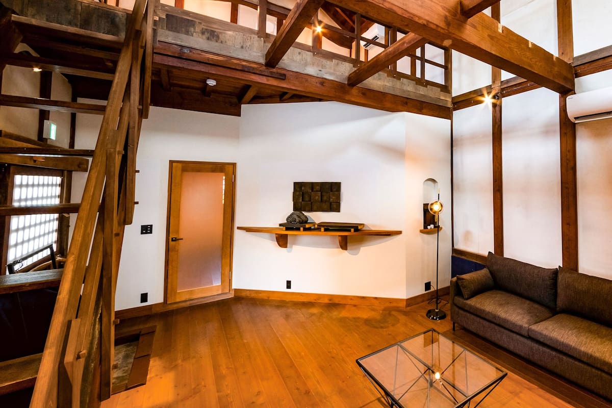 A spacious room with wooden beams, a sofa, a glass coffee table, and a wooden shelf with decorative items.