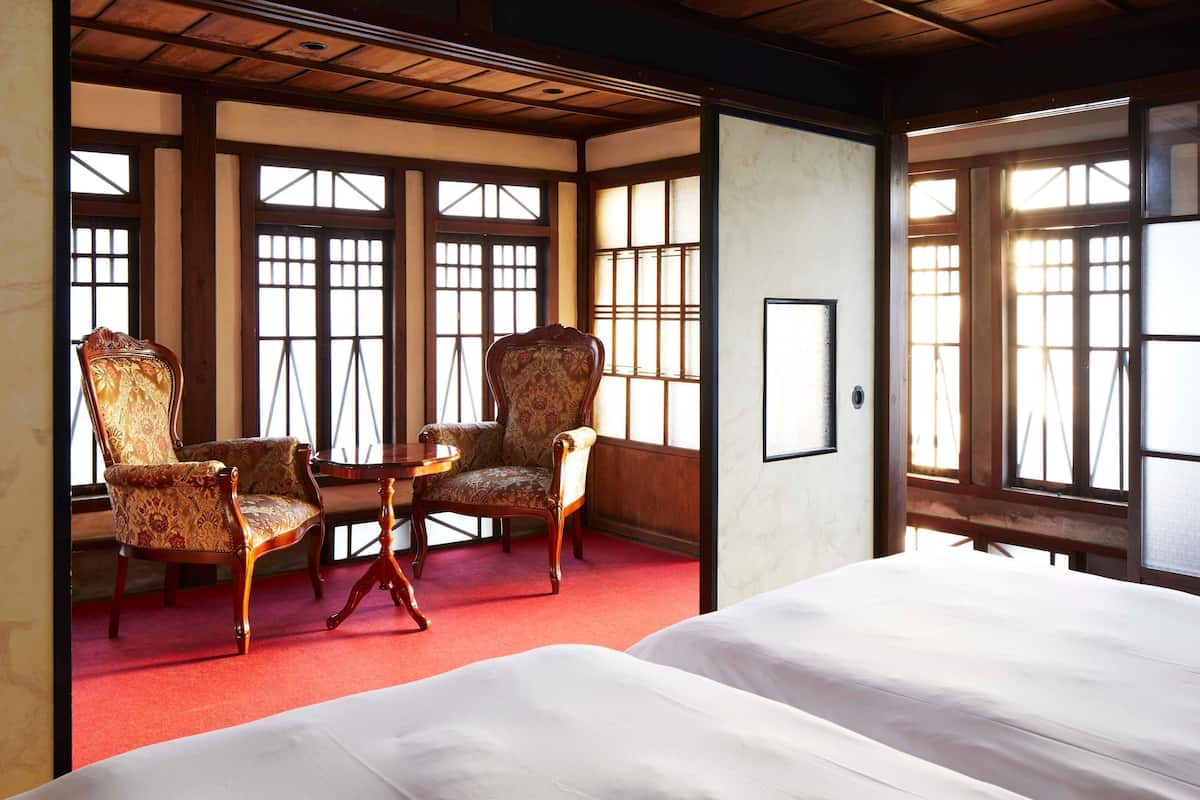 A traditional Japanese room with tatami flooring, a low table, two chairs, and a bed.