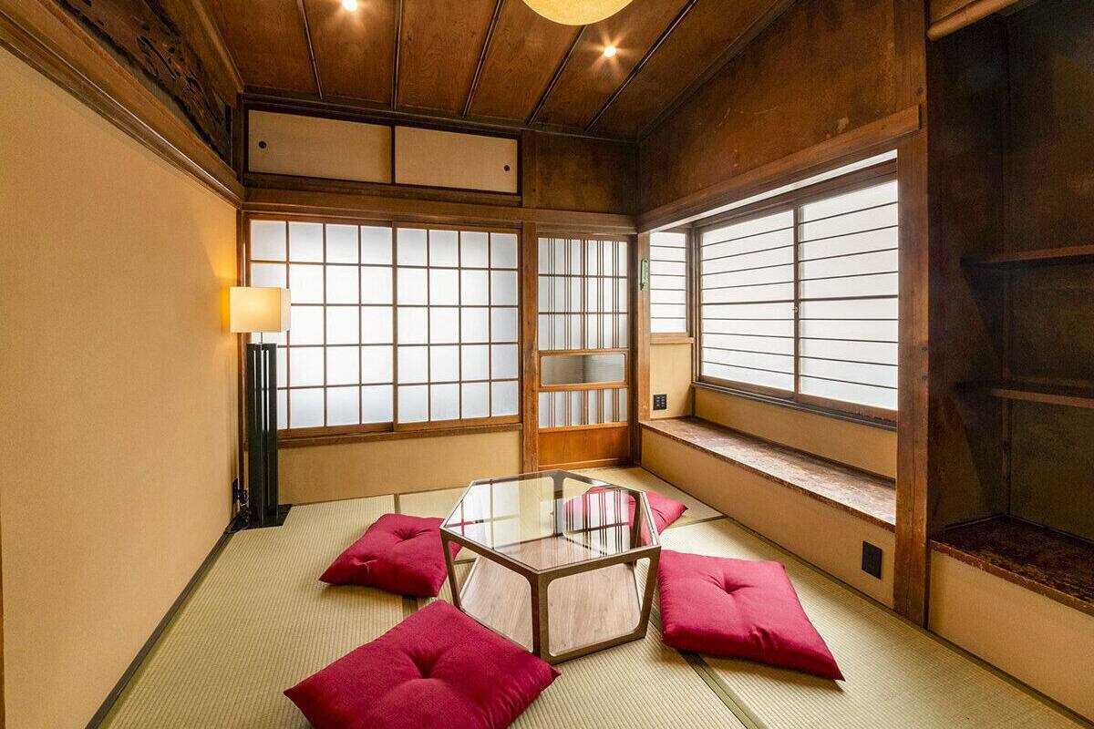 A traditional Japanese room with tatami flooring, a glass table, red cushions, sliding doors, and a lamp.