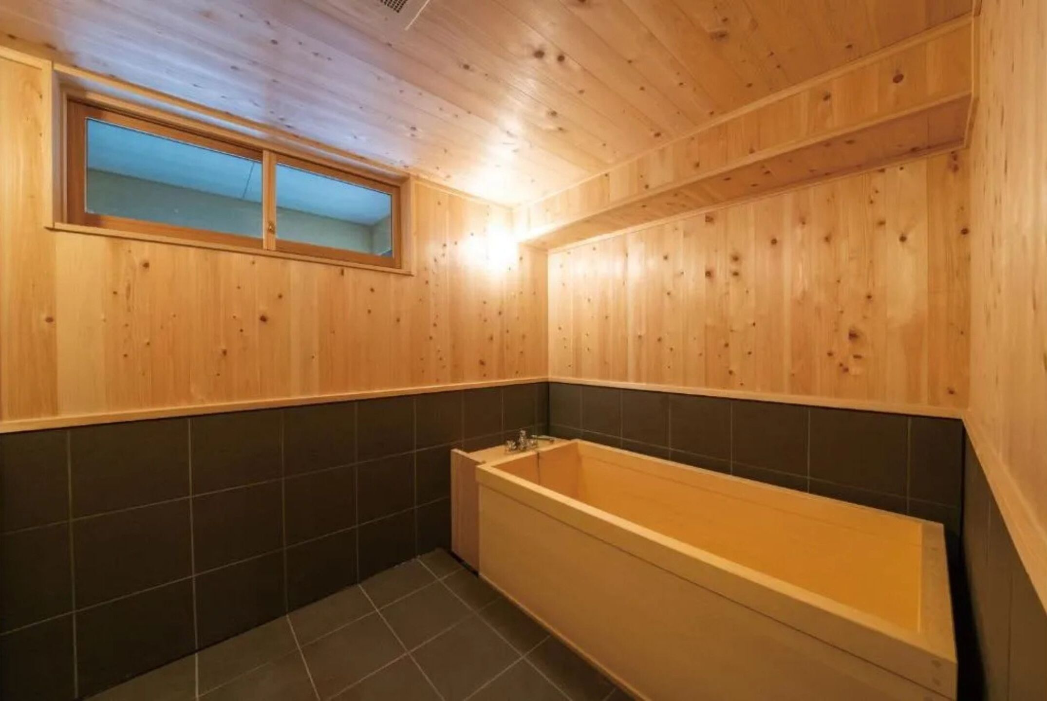 A wooden-paneled bathroom with a bathtub, a window, and dark tiled walls.