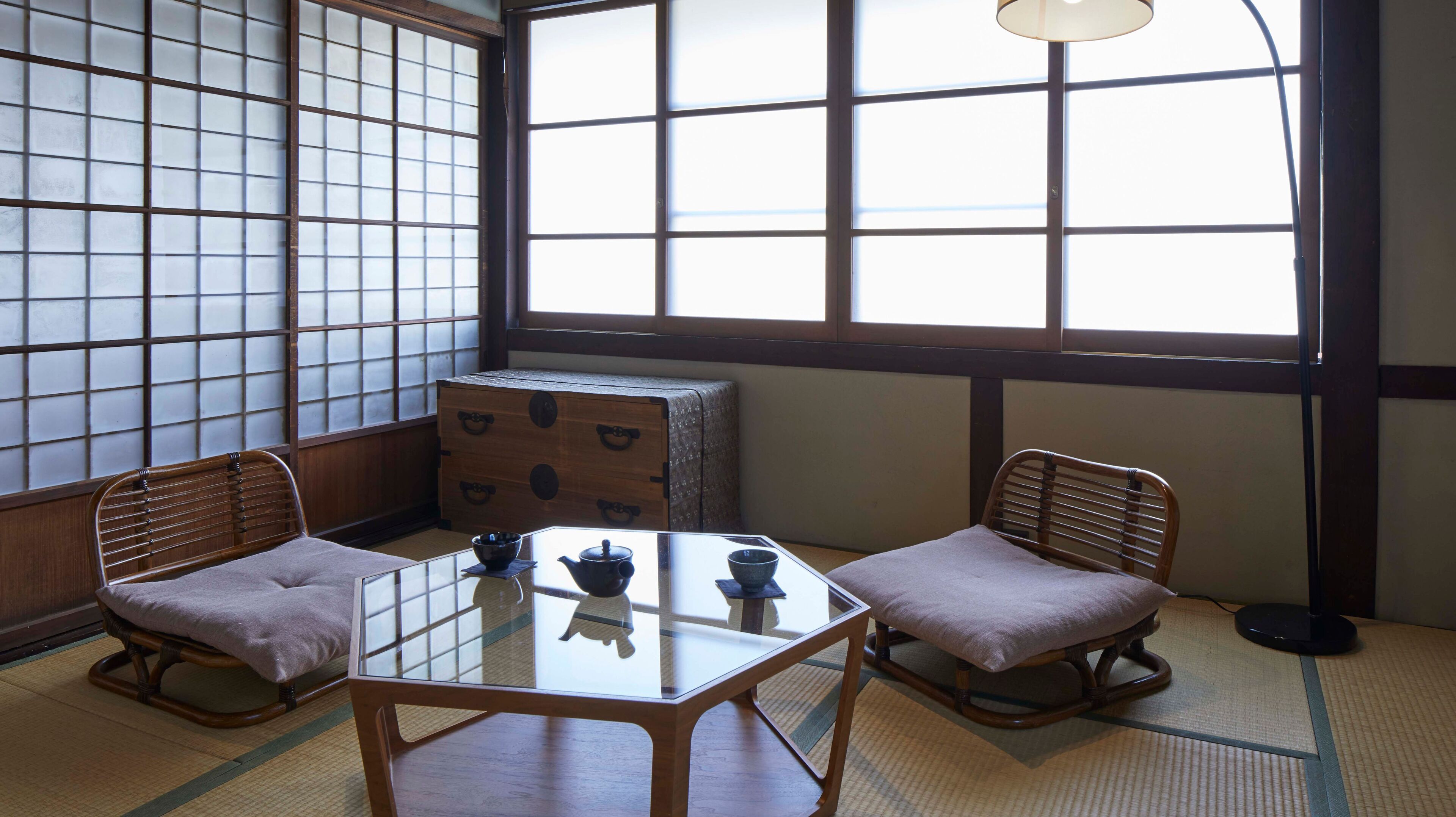 Ett traditionellt japanskt rum med tatamimattor på golvet, ett lågt bord med en glasskiva, två vadderade stolar och en byrå i trä.