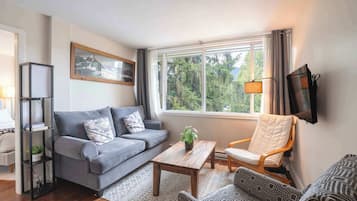 Ein Wohnzimmer mit grauem Sofa, einem Holztisch, einem gemusterten Sessel, einem Bücherregal und einem an der Wand montierten Fernseher.