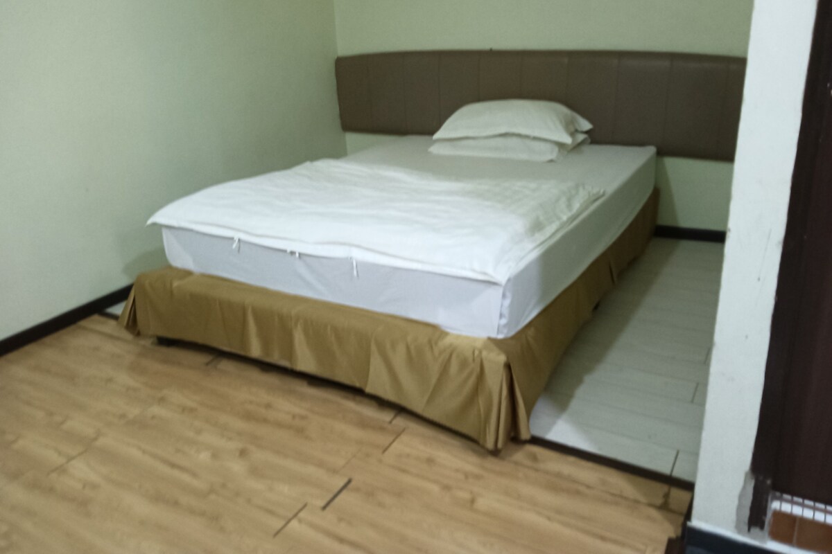 Una cama individual con colchón blanco y falda marrón en una habitación con paredes de colores claros y suelo de madera.