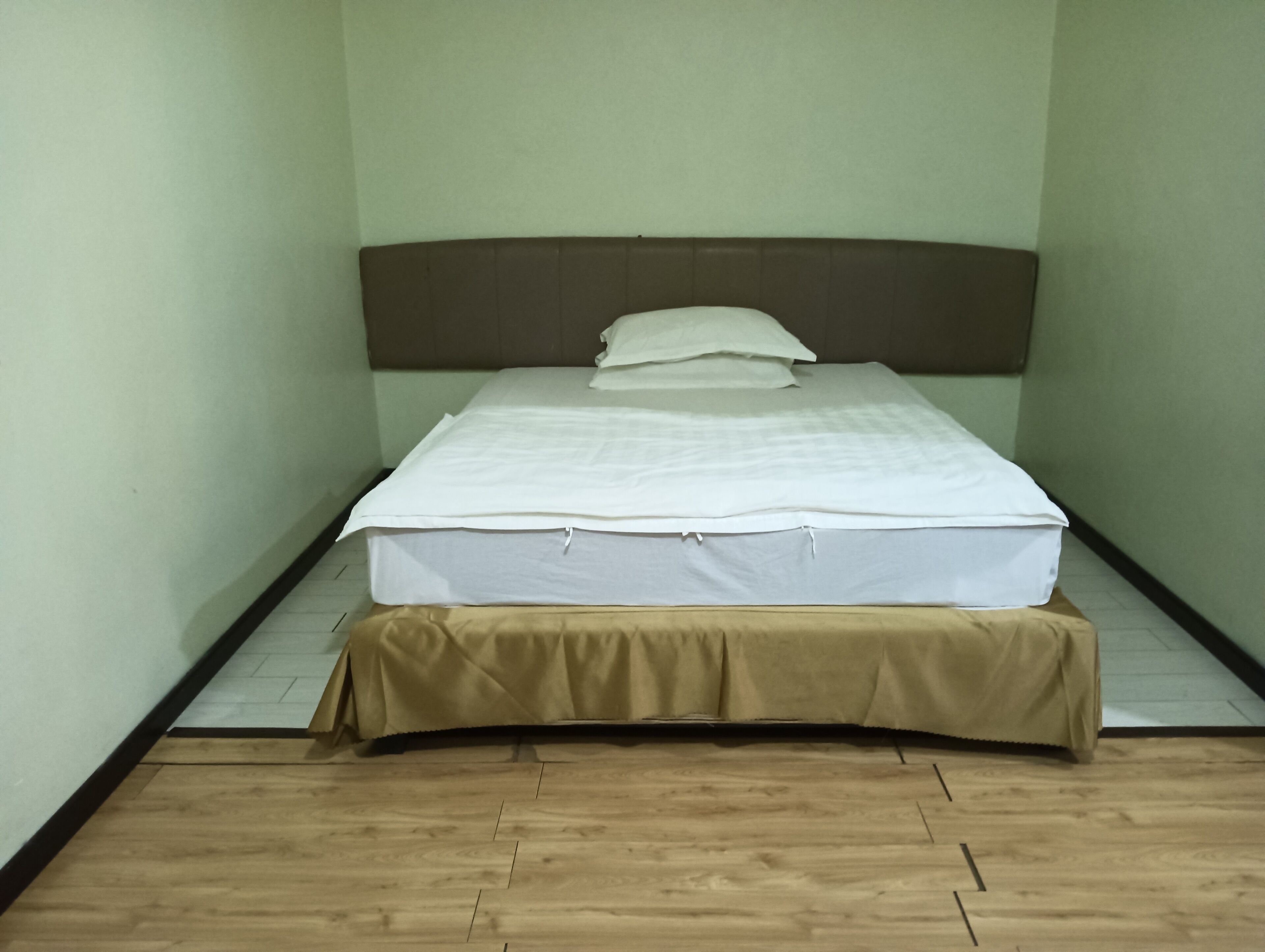Een eenpersoonsbed met een witte matras en een bruin hoofdeinde in een kamer met lichtgroene muren en een houten vloer.