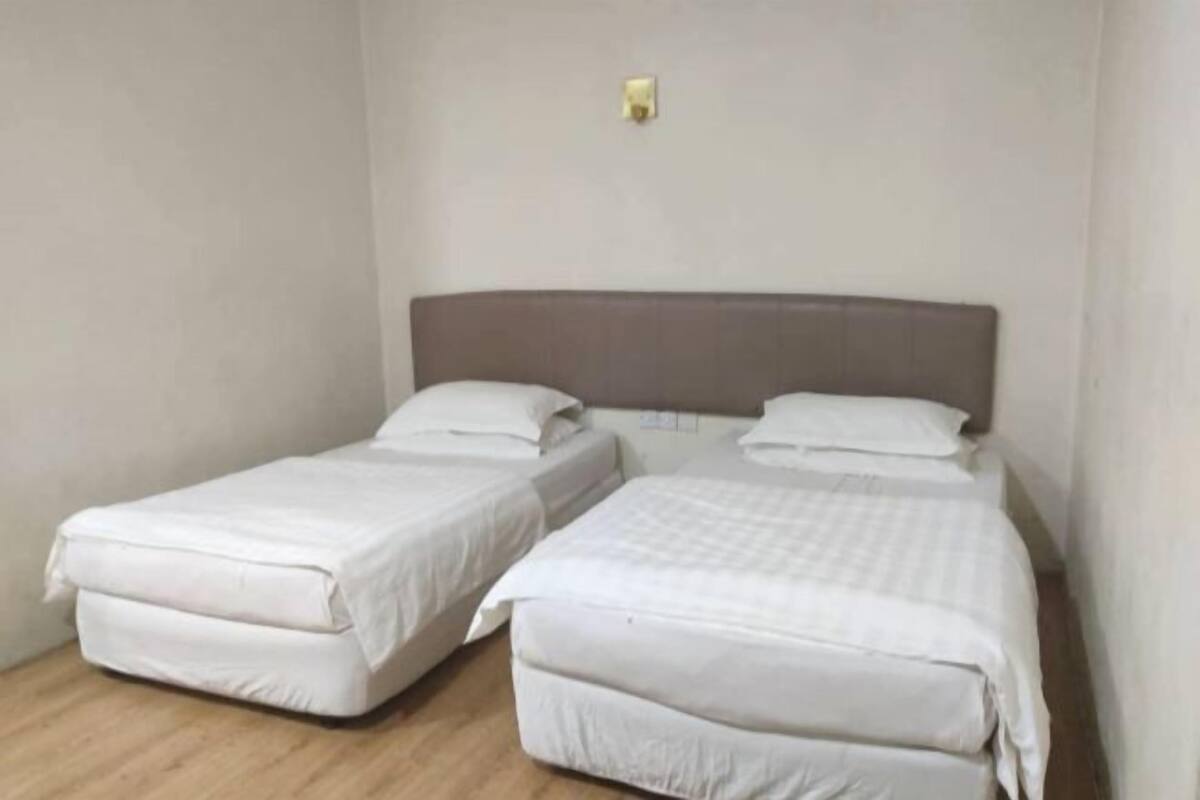 A room with two single beds, a wooden floor, and a plain wall with a light switch.