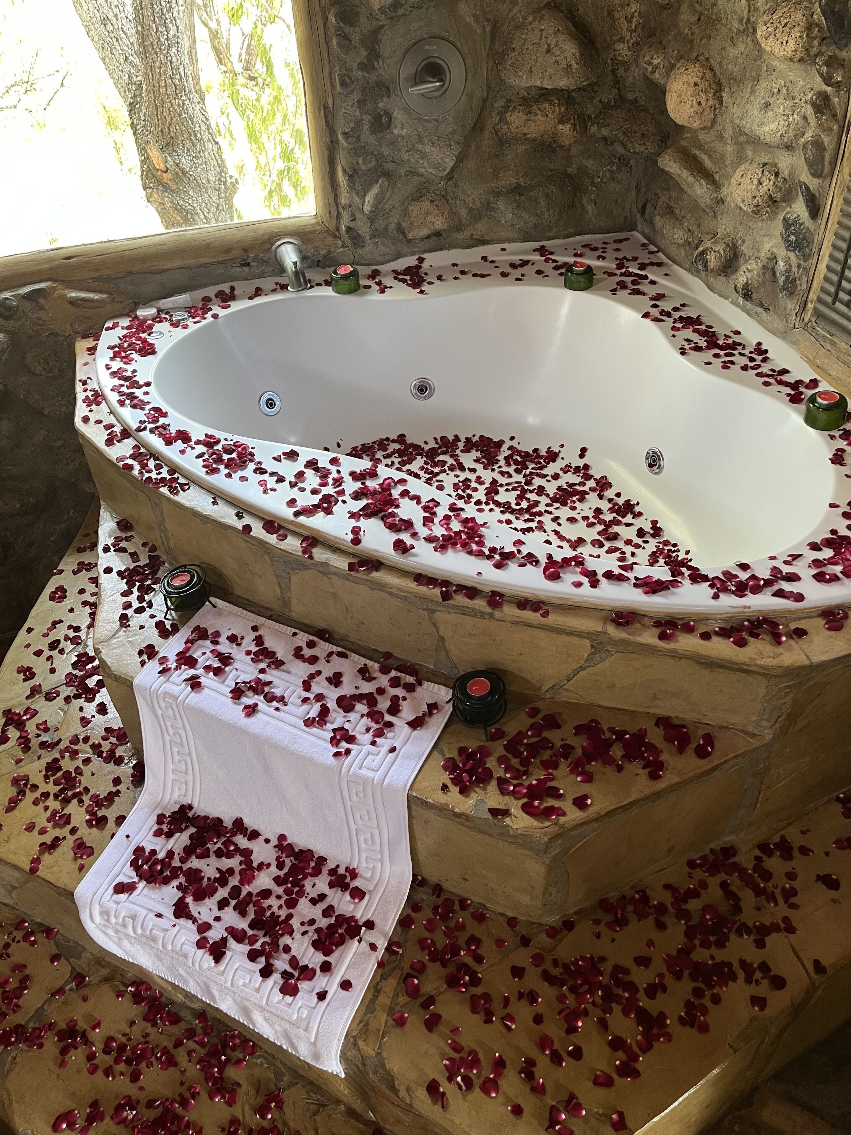A stone-floored bathroom with a freestanding bathtub filled with rose petals and candles.