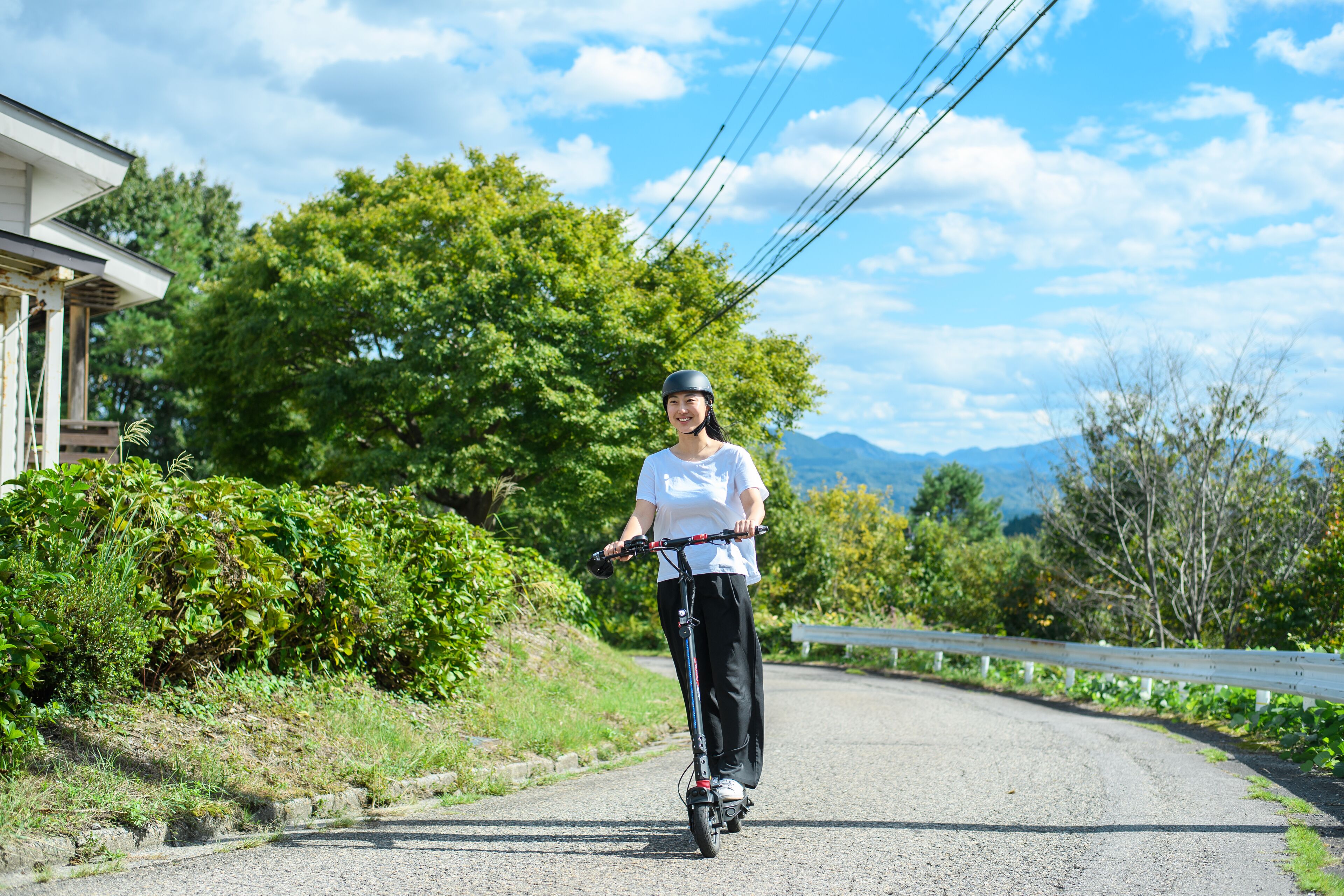 Una persona circulando en patinete eléctrico por un camino rural, con vegetación y una casa a la izquierda.