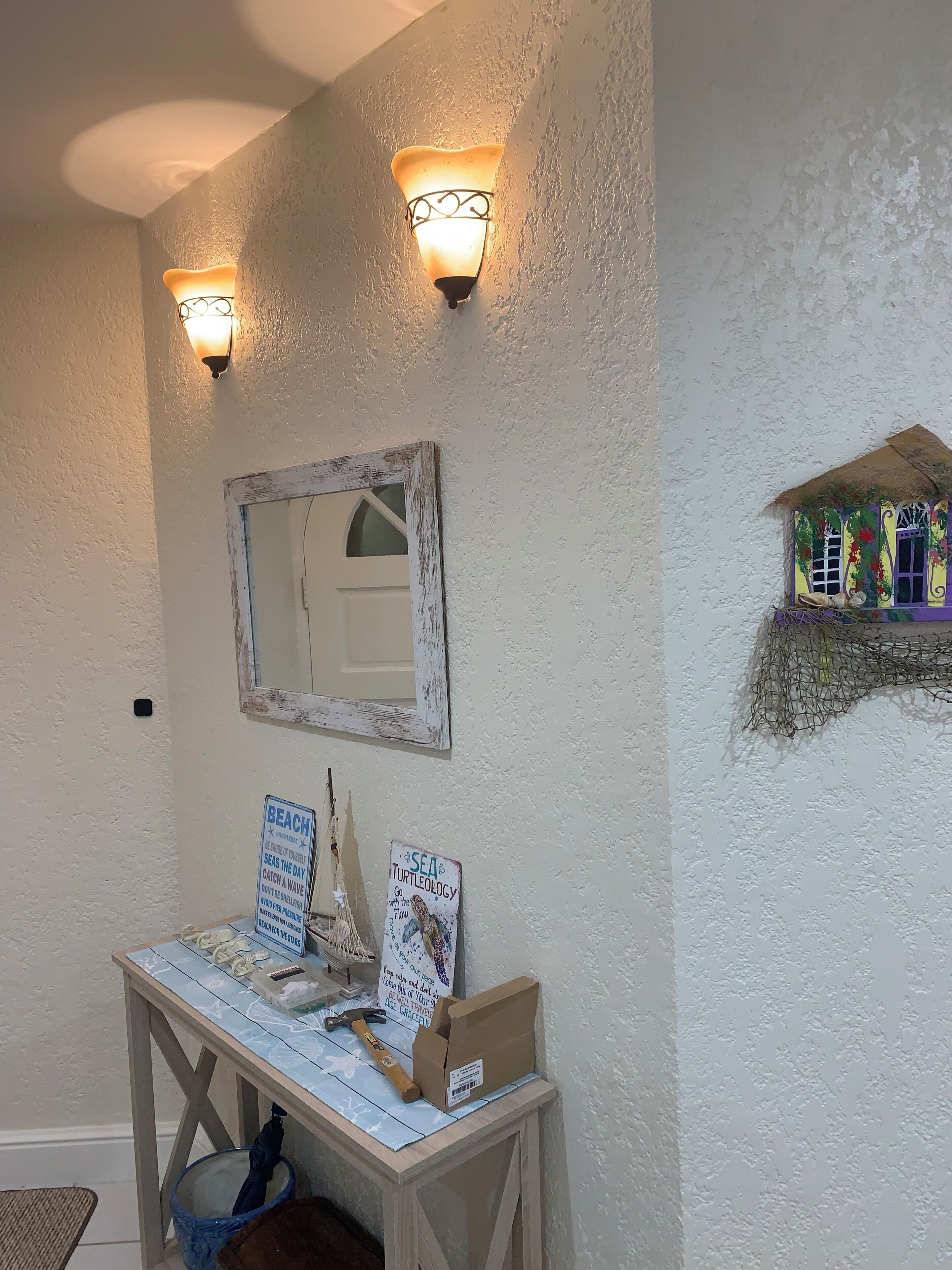 A bathroom with a mirror, two wall-mounted lights, a small table with a towel, a sign, and a decorative item on the wall.