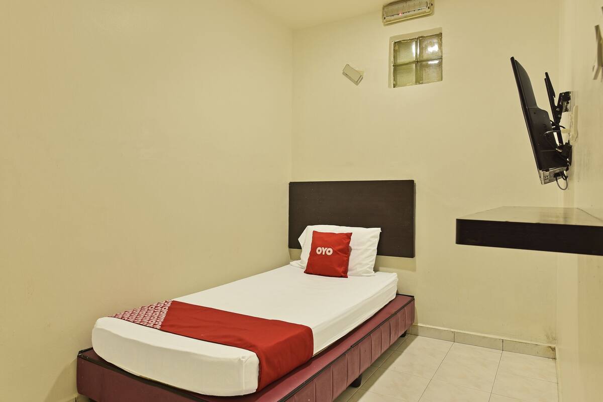 A small, single-bed room with a red and white bedspread, a red pillow, a dark headboard, a small window, and a wall-mounted TV.