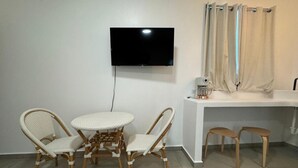 Ein Zimmer mit einem Wand-Fernseher, einem runden Esstisch mit Korbstühlen, einer weißen Kommode mit Kaffeemaschine und zwei Holzschemeln.