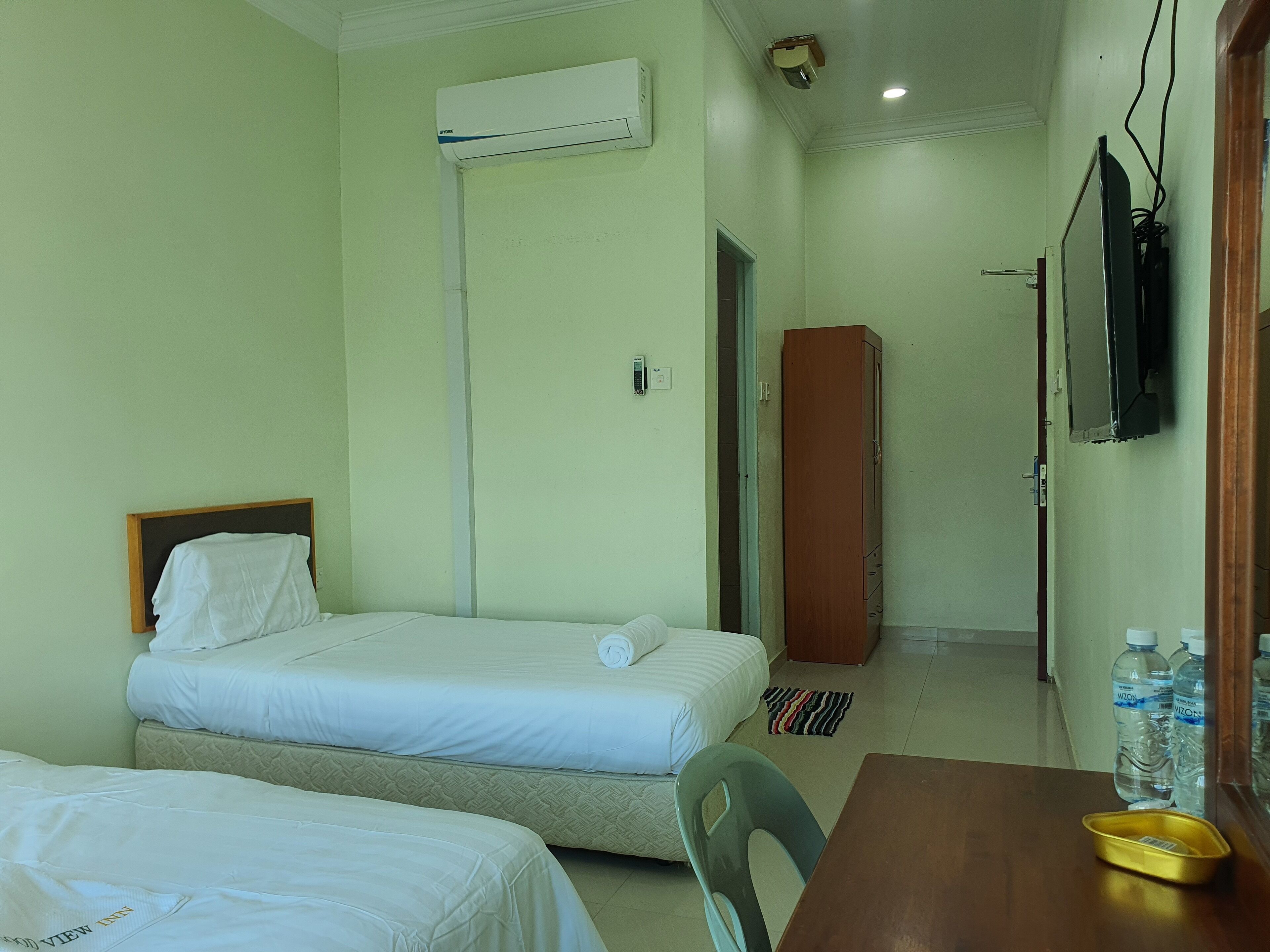 A hotel room with two beds, a wooden wardrobe, a television, and a table with water bottles.