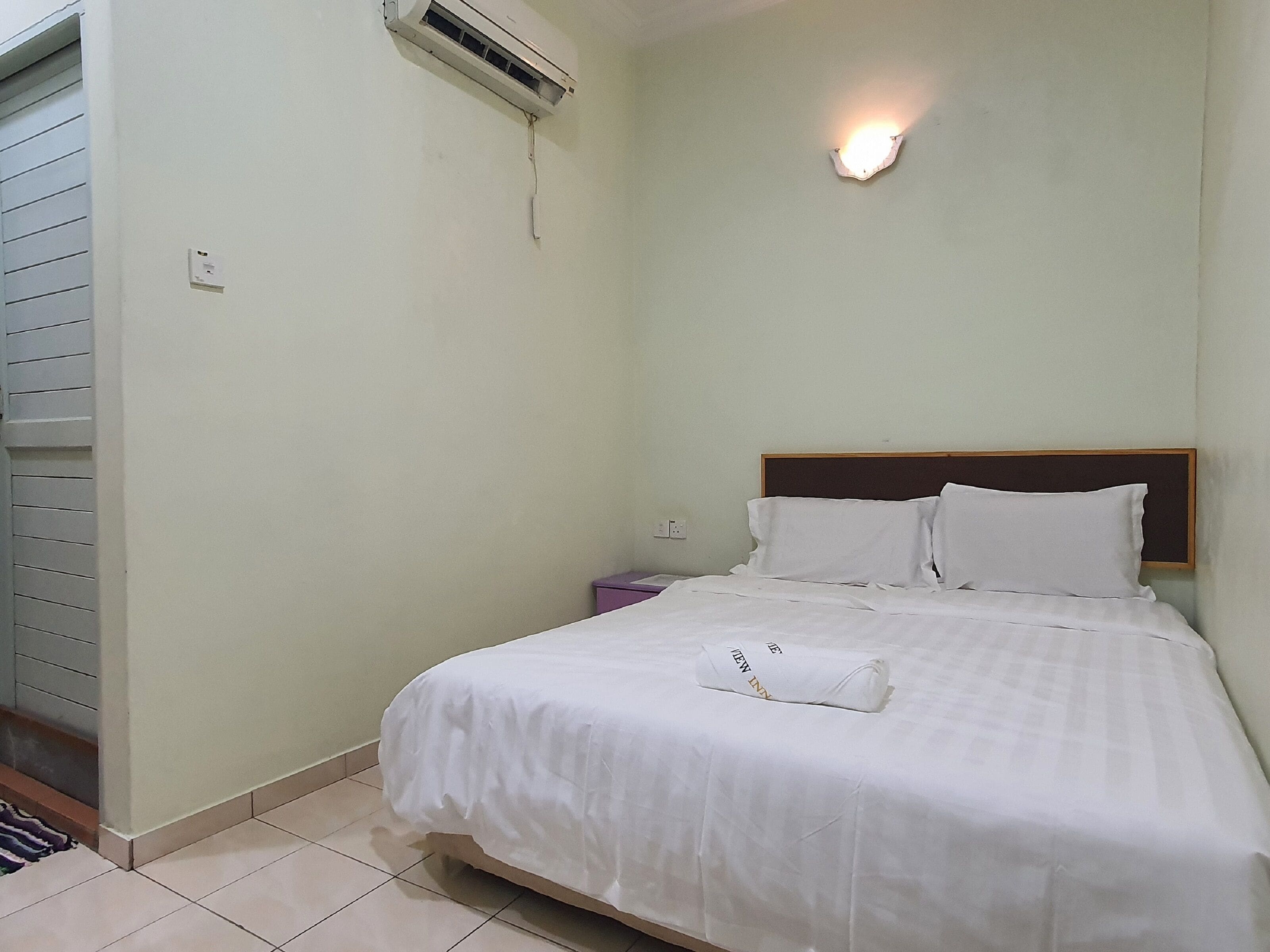 A neatly made bed with white linens, a wooden headboard, a bedside table with a purple lamp, and a wall-mounted air conditioner.