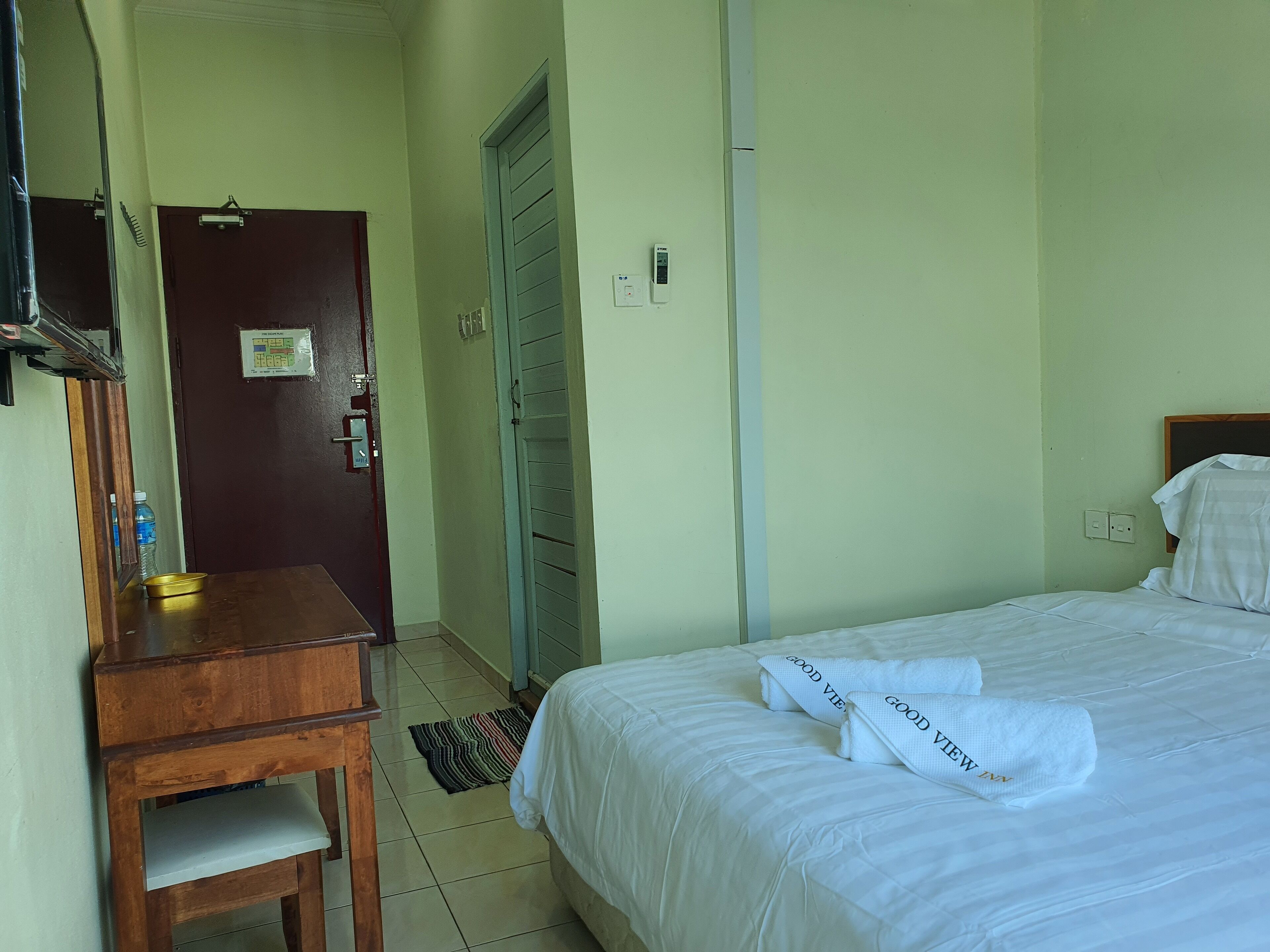A hotel room with a bed, a wooden desk, a chair, a door, and a wall-mounted light switch.