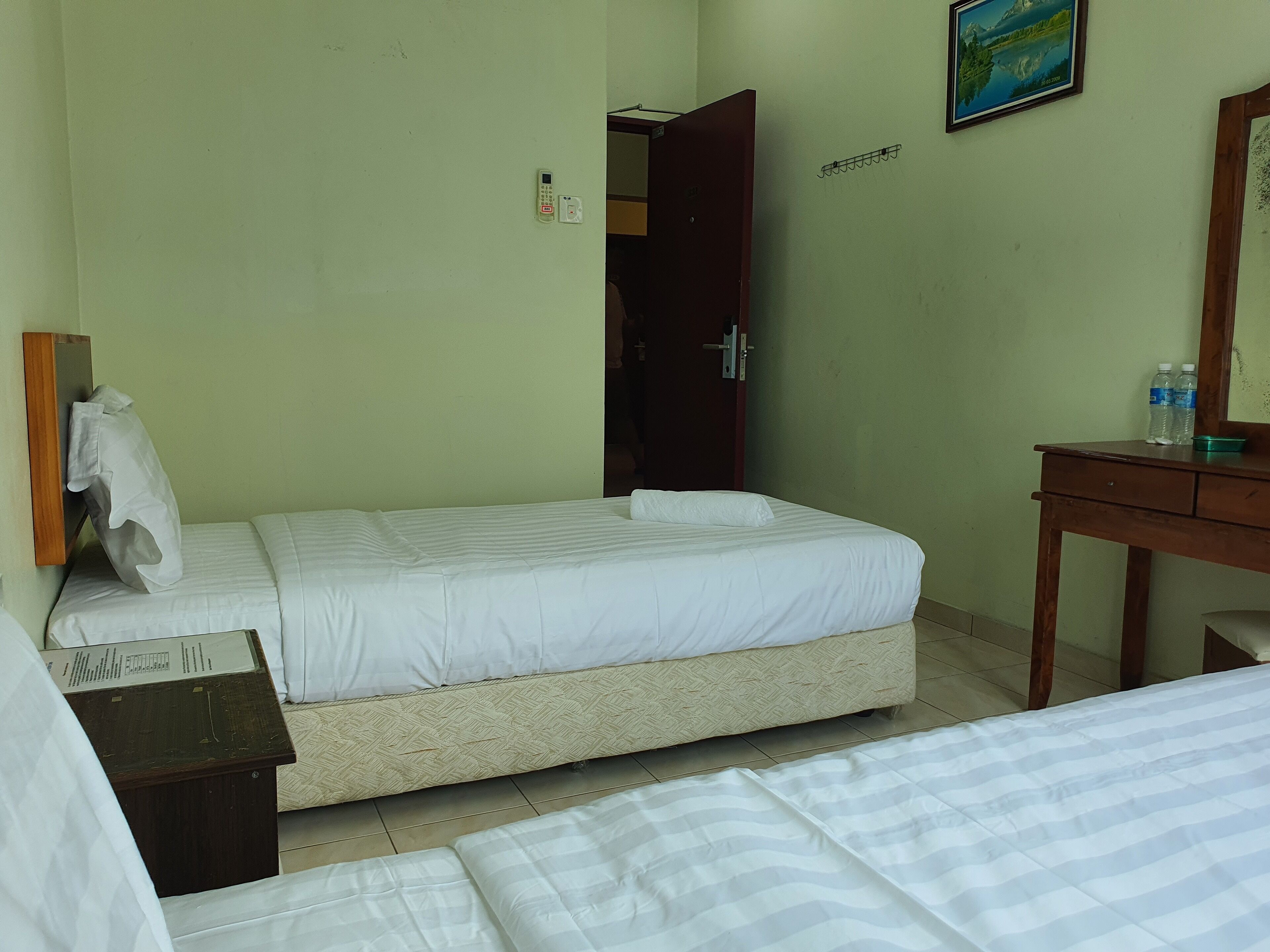 A hotel room with a bed, a wooden desk, a mirror, a door, and a wall-mounted telephone.