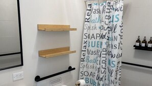 A modern bathroom with a white toilet, a wooden vanity, a white sink, and a shower curtain with text. There are wooden shelves above the toilet and a small shelf with bottles on the wall.