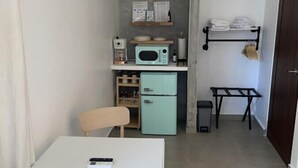 A compact kitchen with a white table, a wooden chair, a teal refrigerator, a microwave, and a coffee maker.