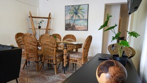Un área de comedor con una mesa redonda y seis sillas de mimbre, un cuadro de una escena de playa y una planta en maceta sobre una mesa negra.