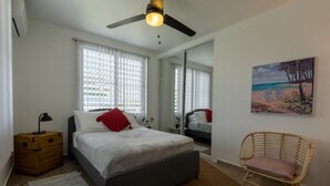 A bedroom with a bed, a ceiling fan, a wooden chest of drawers, a wicker chair, and a painting of a beach scene.