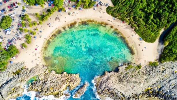 An aerial view of a beach with clear turquoise waters, rocky cliffs, and a sandy beach area with umbrellas and people.