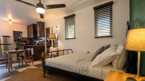 A compact bedroom with a bed, bedside table, and lamp. A kitchenette with a fridge and stove is visible in the background. - Stylish Studio at El Flamingo Beach Club (Tierras Nuevas Poniente)